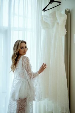 Gelinler günaydın. Nazik ve alımlı, dantel cüppeli sarışın zarif gelinliğin yanında duruyor. Uzun dalgalı saçları var, kameraya bakıyor. İpek bornoz ve iç çamaşırlı genç kadın..