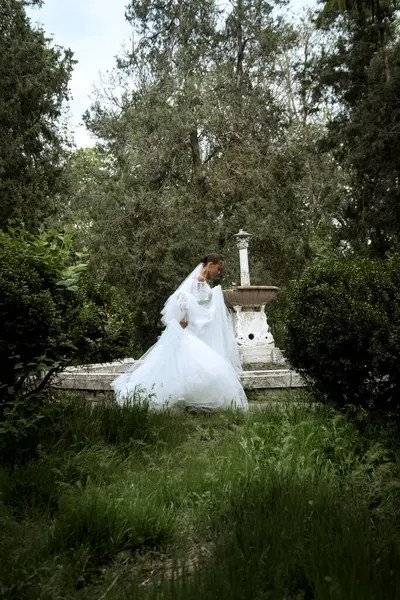 Ağaçların ve eski çeşmelerin arka planında uzun peçeli güzel bir gelin. Uzun beyaz tüylü elbiseli esmer kız gülümsüyor ve seviniyor. Peçe rüzgarda dalgalanıyor.