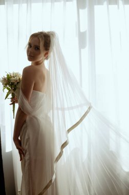 Camın yanında elinde zambak buketiyle uzun beyaz bir elbise içinde duran, gür bir duvağı olan sarışın gelin. Minimalist iç mekan, otel ya da ev. Uzun elbiseli ve uzun saçlı gelin.