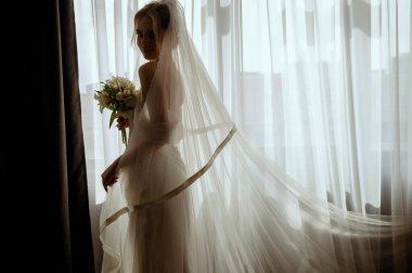 Sarisin gelinin silueti, duvagli, uzun beyaz bir elbiseyle, pencerenin yaninda bir buket zambakla. Minimalist iç mekan, otel ya da ev. Uzun elbiseli ve uzun saçlı gelin.