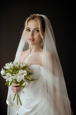 Camın yanında beyaz bir elbise ve bir buket zambak olan sarışın bir gelinin portresi. Minimalist iç mekan, otel ya da ev. Makyajlı gelin ve koyu bir arka planda yüksek saç stili.