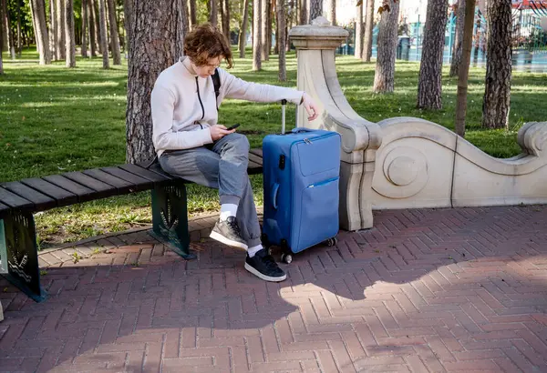 Genç erkek turist, elinde telefon ve mavi bavulla parkta oturuyor. Telefonu elinde tutuyor ve ona bakıyor. açık bej kazaklı ve gri pantolonlu. Avrupa kentinde turizm, Avrupa 'da turizm.