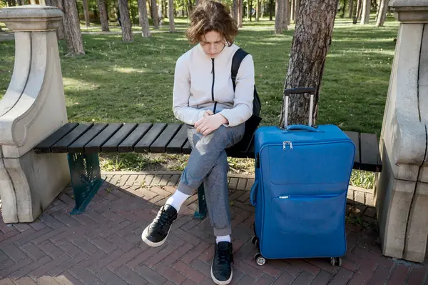 Genç erkek turist, elinde telefon ve mavi bavulla parkta oturuyor. Telefonu elinde tutuyor ve ona bakıyor. açık bej kazak ve gri pantolonla güneşli havada, Avrupa 'da turizm