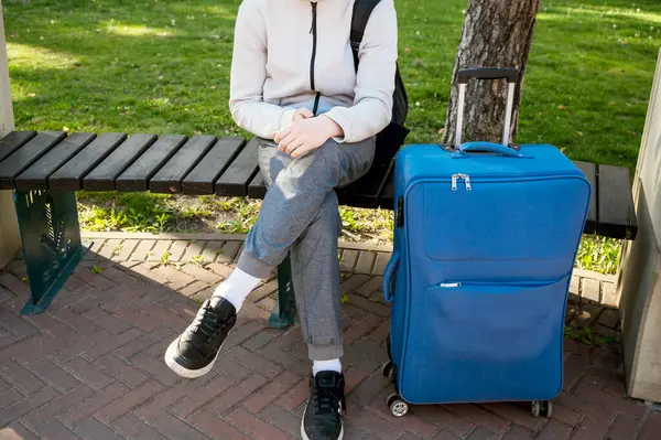 Parkta elinde telefon ve mavi bir bavulla oturan bir turist. Telefonu ellerinin arasında tutuyor ve ona bakıyor. Açık bej kazaklı ve gri pantolonlu. Avrupa 'da Turizm