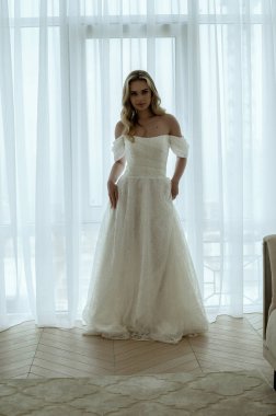 Sarışın gelin, dantelli bir gelinlikle pencerenin önünde duruyor. Büyük bir pencerenin arkasında. Gelinlerin sabahı, düğün günü konsepti. Minimalist bir iç mekanda, parlak bir odada.