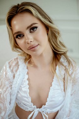 Dantel bornoz ve ipek iç çamaşırı giymiş genç bir sarışının yakın çekimi. Gelinlerin sabahki konsepti, gelin hazırlıkları, dinlenme, makyaj, kişisel bakım. Kızın dalgalı saçları ve makyajı var..