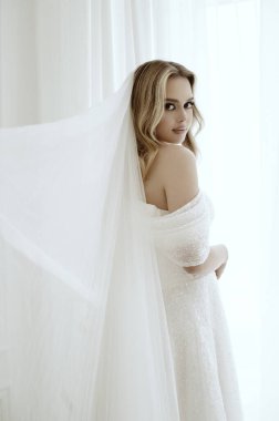 Beyaz elbiseli, lüks duvağı olan sarışın gelin parlak odanın penceresinin yanında duruyor. Düğün günü konsepti, aşk, düğün aksesuarları ve kıyafetler. Sarışın, minimalist iç mekanda kameraya bakıyor.