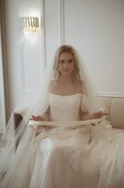 Duvak takmış beyaz elbiseli gelin beyaz odada oturuyor. Düğün günü konsepti, aşk, düğün aksesuarları ve kıyafetler. Minimalist iç mekanda kameraya bakan sarışın.