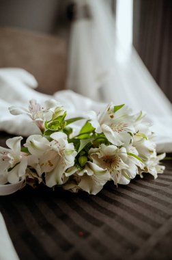 Düğün detayları. Gelin aksesuarları. Ön plandaki çiçekler keskin odak noktasında, arka plan bulanık. Zarif bir gelinlik, arka planda karanlık bir yatakta uzanır. Lüks bir duvak.