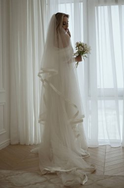 Lüks, uzun ve şık elbiseli gelin pencerenin arkasına poz veriyor. Gelin silueti ve zambak buketi olan kocaman bir pencere. Gelin damadı bekliyor..