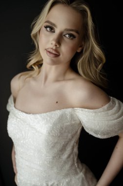 Zarif bir gelinliğin portresi, kapalı çekimler sırasında bir modelle birlikte minimalist bir ortamda sergileniyor. Sarışın gelin, pencerenin yanındaki karanlık iç kısma karşı.. 