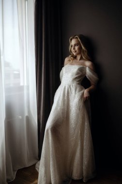 Zarif bir gelin uzun, beyaz bir gelinlik içinde poz verir. Pencerenin yakınındaki koyu minimalist bir iç mekanın arka planında tam bir büyüme var. Karanlık arka plan onun klasik güzelliğini vurguluyor.