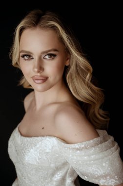 Genç, dalgalı saçlı, sarışın bir kadın. Uzun saçlı, güzel bir manken. Mükemmel makyajlı ve şekilli bir sarışın. Omuzları açık beyaz bir elbise içinde..