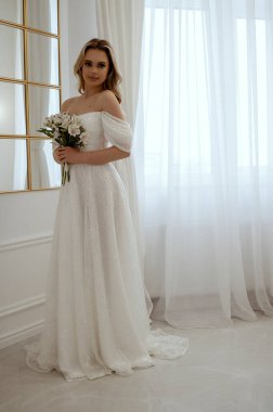 Pencere kenarında duran bir buket beyaz çiçekli gelin. Dalgalı bir gelinlik içindeki sarışın, zarafet ve güzellik saçıyor. Penceresi ve aynası olan minimalist bir iç mekanda. 