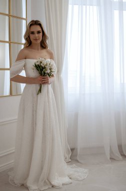 Pencere kenarında duran bir buket beyaz çiçekli gelin. Dalgalı bir gelinlik içindeki sarışın, zarafet ve güzellik saçıyor. Penceresi ve aynası olan minimalist bir iç mekanda