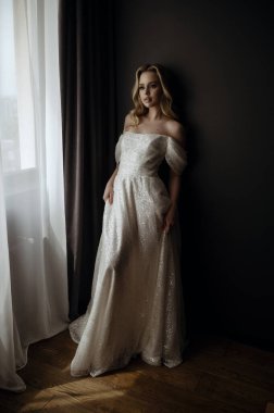 Zarif gelinlik, kapalı çekimler sırasında şık pozlar veren bir modelle minimalist bir ortamda sergileniyor. Zarif bir gelin uzun, beyaz bir gelinlik içinde poz verir. Yüksek kalite fotoğraf