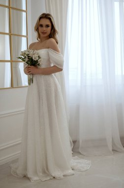 Gelin, pencerede beyaz bir buket çiçekle dimdik ayakta duruyor. Dalgalı bir gelinlik içindeki sarışın, zarafet ve güzellik saçıyor. Penceresi ve aynası olan minimalist bir iç mekanda.