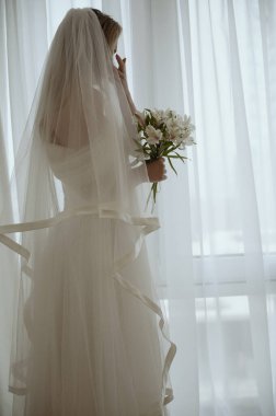 Lüks, uzun ve şık elbiseli gelin pencereye poz veriyor. Gelin silueti ve zambak buketi olan kocaman bir pencere. Gelin damadı bekliyor ve gözyaşlarını siliyor