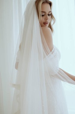 Beyaz elbiseli zarif sarışın gelinin portresi. Duvağı yakın çekimde poz veriyor. Gelin portresi, düğün makyajı, saç stili, şık bir gelin. Peçeli güzel gelin.