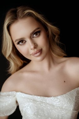 Genç bir portre, dalgalı saçlı sarışın bir kadın. Uzun saç stili olan güzel bir model. Mükemmel makyajlı sarışın. Omuzları açık beyaz bir elbise içinde. Karanlık bir arkaplanda kameraya bak.