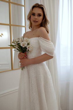 Penceresinde bir buket beyaz çiçekle sarışın bir gelinin portresi. Dalgalı bir gelinlik içindeki sarışın, zarafet ve güzellik saçıyor. Penceresi ve aynası olan minimalist bir iç mekanda