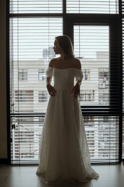 Kocaman bir pencereye karşı bir gelin silueti. Minimalist bir iç mekanda şık bir modelle sunulmuş zarif bir gelinlik. Sarışın bir gelin karanlık bir iç mekâna karşı.