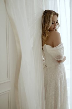 Beyaz elbiseli, lüks duvağı olan sarışın gelin parlak odanın penceresinin yanında duruyor. Düğün günü konsepti, aşk, düğün aksesuarları ve kıyafetler. 
