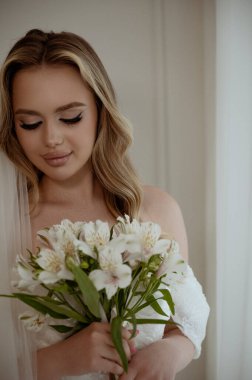Sarışın bir gelinin portresi, gür bir peçesi, beyaz bir elbise ve bir buket zambak. Beyaz arka planda yakın plan. Makyajlı ve dalgalı uzun saçlı gelin