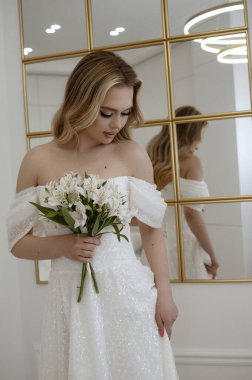 Sarışın bir gelinin portresi bir buket beyaz çiçekle aynanın karşısında. Sarışın, beyaz zambaklar tutan, dalgalı bir gelinlik içinde poz veriyor. İçi aydınlık, büyük bir aynaya karşı.
