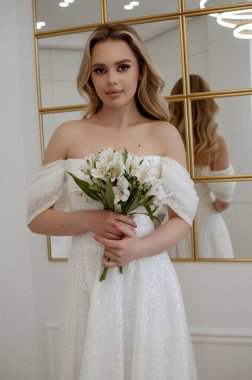 Sarışın bir gelinin portresi, bir demet beyaz çiçekle bir aynanın arka planında. Sarışın, akıcı bir gelinlik giymiş, arka planda bir ayna var, kameraya bakıyor.