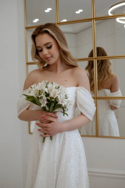 Penceresinde bir buket beyaz çiçekle sarışın bir gelinin portresi. Dalgalı gelinlikli sarışın. Bir pencerenin arka planına karşı hafif bir iç mekânda. Uzun saçlı.