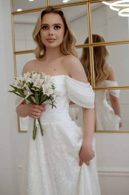 Sarışın bir gelinin portresi, bir demet beyaz çiçekle bir aynanın arka planında. Dalgalı gelinlikli sarışın. İçi aydınlık, mutlu bir şekilde gülümsüyor.