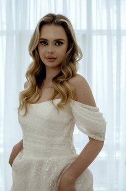 Göz kamaştırıcı bir gelin, minimalist bir iç mekanda beyaz arka planda dalgalı bir gelinlikle zarif bir poz verir. Zarafet ve güzellik saçar