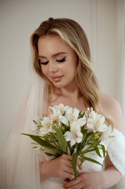 Sarışın bir gelinin portresi, gür bir peçesi, beyaz bir elbise ve bir buket zambak. Beyaz arka planda yakın plan. Makyajlı, dalgalı uzun saçlı, gülümseyen bir gelin.