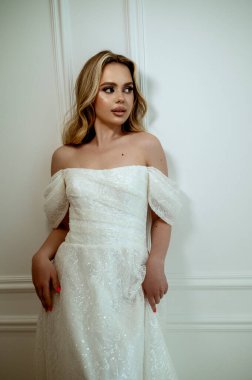 Göz kamaştırıcı bir gelin, minimalist bir iç mekanda beyaz arka planda dalgalı bir gelinlikle zarif bir poz verir. Zarafet ve güzellik saçıyor. Yüksek kaliteli fotoğraf.