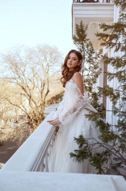 Geleneksel uzun beyaz elbiseli esmer gelin baharda otel balkonunda poz veriyor. Uzun kollu beyaz elbise, korse ve açık omuzlu. Klasik mimarisi olan bir otel. Gelin portresi