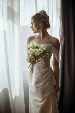 Pencerenin yanında uzun beyaz bir elbise içinde elinde zambak buketiyle güzel sarışın gelin duruyor. Minimalist iç mekan, otel ya da ev. Yan tarafında kesik olan uzun elbiseli gelin.