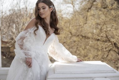 Geleneksel uzun elbiseli esmer gelin baharın başında beyaz otel balkonunda poz veriyor. Uzun kollu beyaz elbise, korse, açık omuzlar. Gelinin portresi, düğün makyajı ve saç stili.