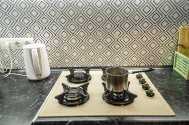 Mutfağın içinde küçük bir tenceresi olan dahili bir gaz sobasının yakın çekimi. Gaz hobisi açık bej, yanında beyaz bir elektrikli su ısıtıcısı var..