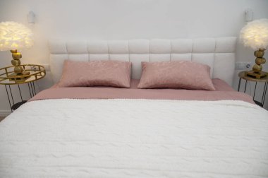 Beyaz yatak başlıklı çift kişilik yatak ve koyu pembe yatak örtüsü ve beyaz battaniye. Yuvarlak metal komodinlerin üzerinde beyaz yuvarlak tüy lambaları var. Yerleşim için uygun. Ön manzara. Yatak odasındaki duvarlar beyaz.