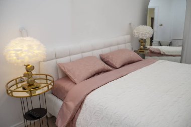 Yatak odasında çift beyaz yatak, koyu pembe çarşaflı ve beyaz battaniyeli. Yatak odasında iki yastık, beyaz tüy lambalı iki komodin. Yandan bir fotoğraf. Düzen için uygun.