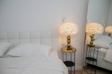 Yatak odasının içinde modern okuma lambası ve yastıklı çift kişilik yatağın yanında. Yuvarlak masa, beyaz tüy lambası, ayna. Konforlu dairede akşam ışığı. Rahat bir otel odası, ışıklandırma ekipmanları..