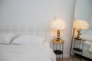 Yatak odasının içinde çift kişilik yatağın yanında modern okuma lambası var. Yuvarlak masa, beyaz tüy lambası, ayna. Işıl ışıl bir dairede akşam ışığı. Rahat bir otel odası, ışıklandırma ekipmanları..