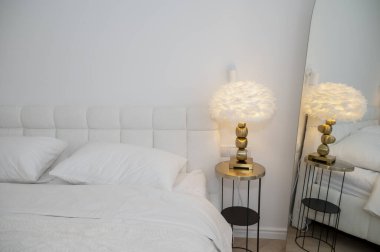 Yatak odasının içinde çift kişilik yatağın yanında modern okuma lambası var. Yuvarlak masa, beyaz tüy lambası, ayna. Işıl ışıl bir dairede akşam ışığı. Rahat bir otel odası, ışıklandırma ekipmanları..