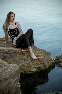Siyah elbiseli ve inci kolyeli genç sarışın denizin arka planında sahilde bir taşın üzerinde oturuyor. Sakin su yüzeyi. moda konsepti, kadın giyimi, barış ve yansıma