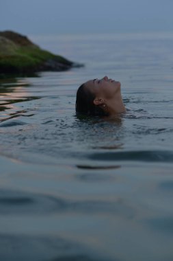 Akşam denizi ve kayaların arkasında genç bir kadının yakın çekimi. Sakin bir denizin arka planında siyah mayo giymiş bronzlaşmış bir kadın. Gözleri kapalı, yüzüyor, saçları ıslak.