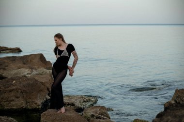 Güzel sarışın, akşam denizinin arka planında kayaların üzerinde poz veriyor. Etrafında kayalar var ve deniz yüzeyi sakin. Siyah, dar giysiler ve şık mücevherlerle. Yalın ayak ayakta