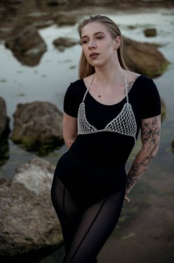 Sarışının yakın planında, akşam denizinin arka planında kayaların üzerinde duruyor. Etrafında kayalar ve sakin bir deniz var. Siyah, dar giysiler ve şık kristal mücevherlerle