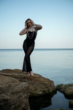 Sarışın, akşam denizinin arka planında poz veriyor, denizin ortasında, büyük bir kayanın üzerinde duruyor, zarif siyah bir takım elbise içinde, boynunda büyük inci mücevherlerle. Sahilde yalınayak durmak