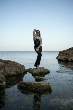 Sarışın, uzun saçlı, akşam denizinin arka planında poz veriyor, büyük bir kayanın üzerinde, denizin ortasında, şık siyah bir takım elbise içinde, boynunda büyük incilerle. Yalın ayak duruyor.